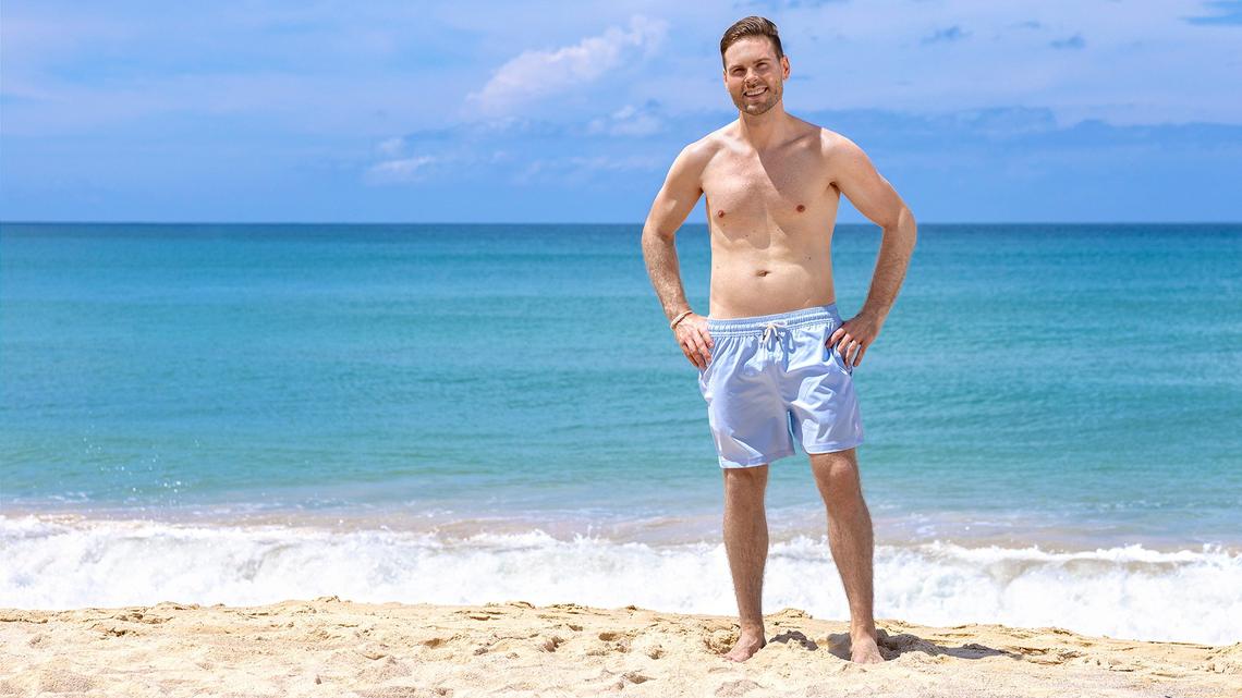 Der Bachelorette-Kandidat Heinz Baumann in der Badehose an einem Strand. Hinter ihm ein ruhiges, blaues Meer.