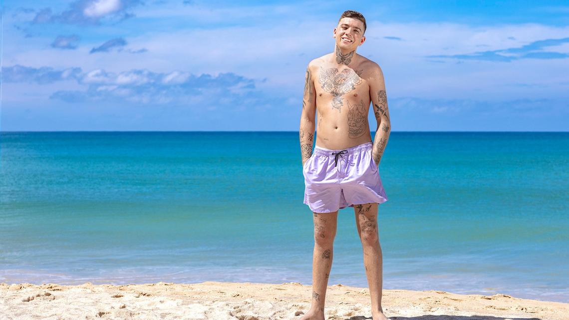Giuliano Hediger trägt lila Badehosen und steht an einem weissen Sandstrand vor einem blauen Meer.