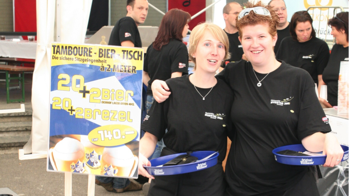 Zwei Helferinnen mit Tablett in schwarzer Kleidung in einem Festzelt draussen.