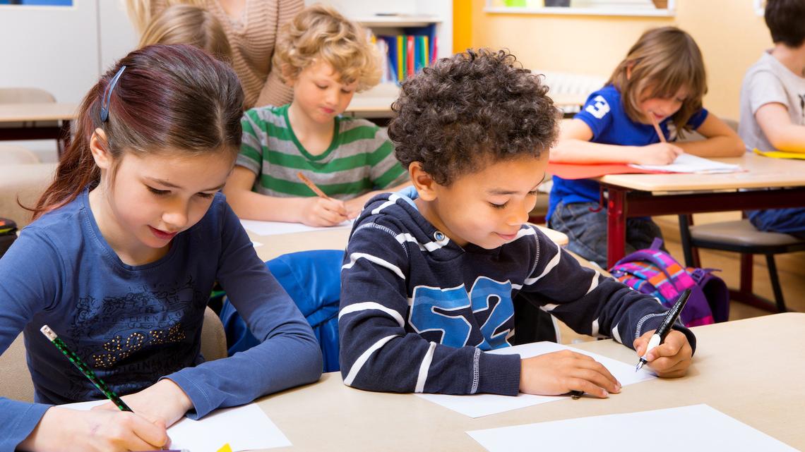 Schulkinder schreiben an ihren Tischen. Ein Junge und ein Mädchen im Vordergrun, im Hintergrund drei Kinder und eine Lehrperson.