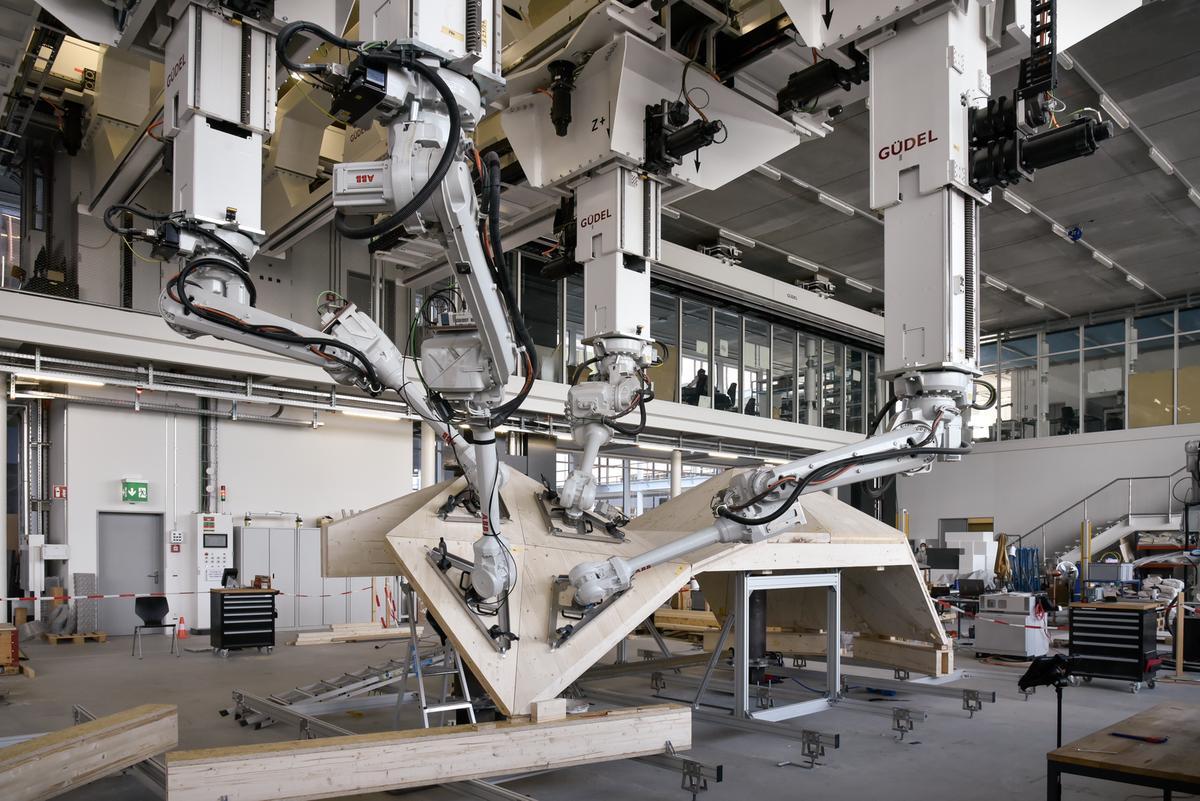 Vier Roboterarme fügen in einer Fabrikhalle Holzplatten zusammen.
